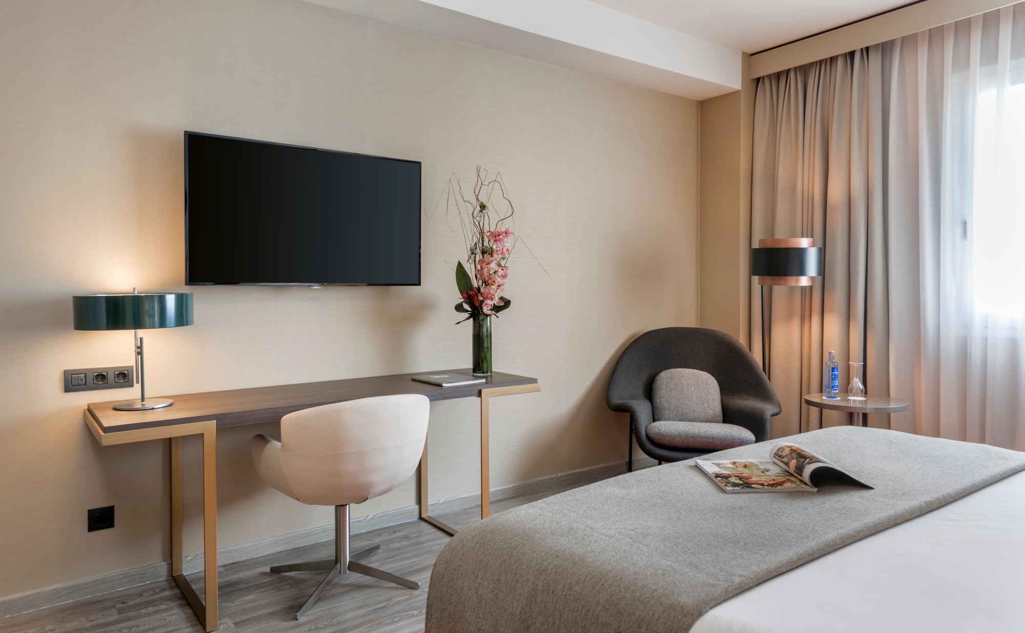 A guest room with a wall-mounted television above a slim desk, a white swivel chair, a dark upholstered armchair, a floor lamp and a bed dressed with a grey runner.