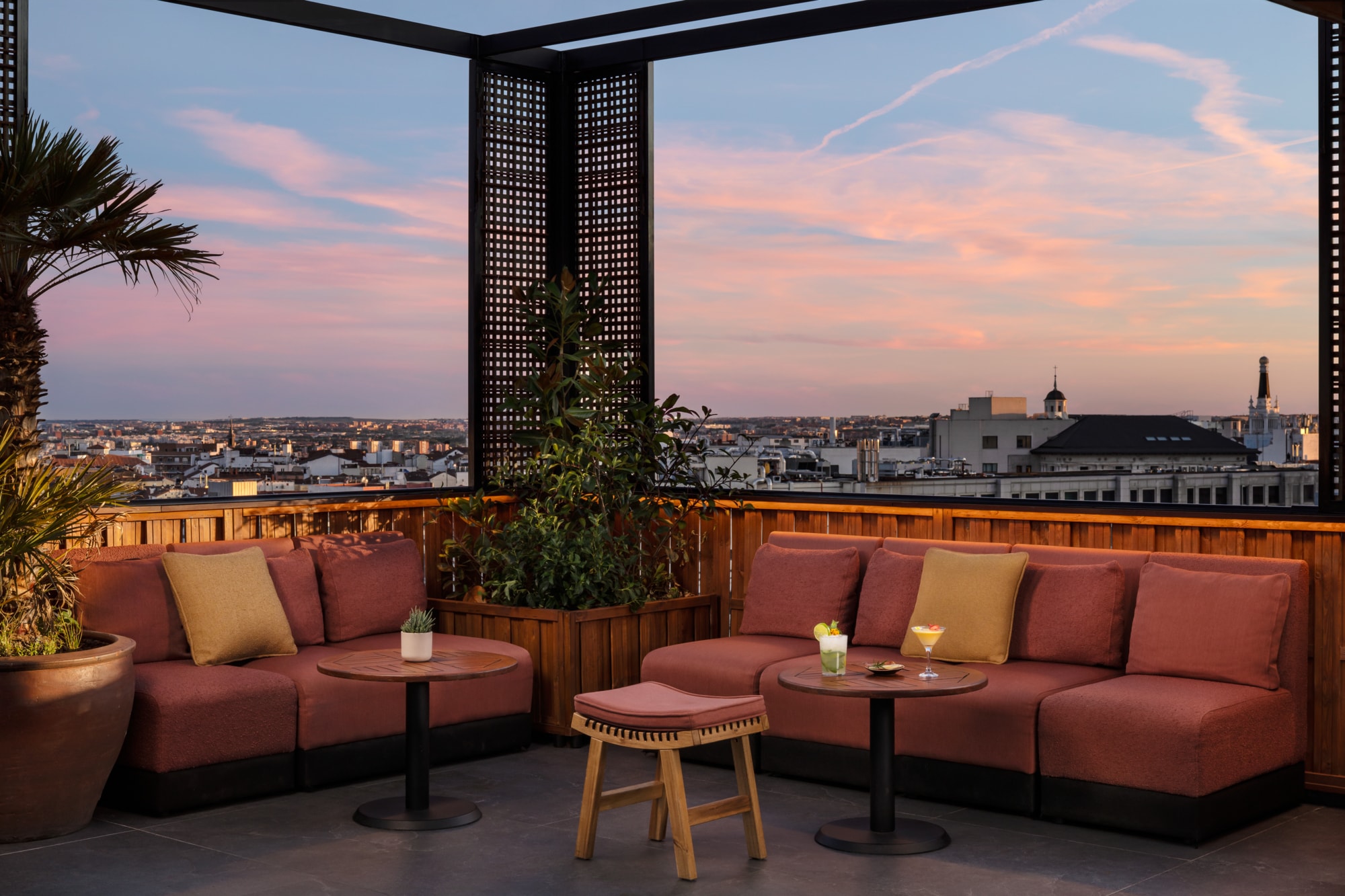 Rooftop terrace at dusk with coral-cushioned banquettes, low round tables holding two cocktails, potted plants and Madrid rooftops beyond the wooden railing.