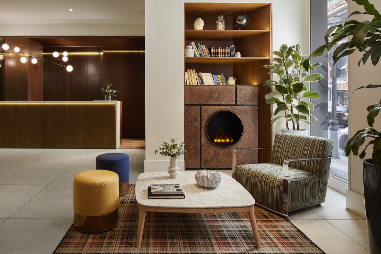 The lobby lounge with a marble coffee table, yellow and navy poufs, a green armchair and a fireplace set into a bookcase wall.