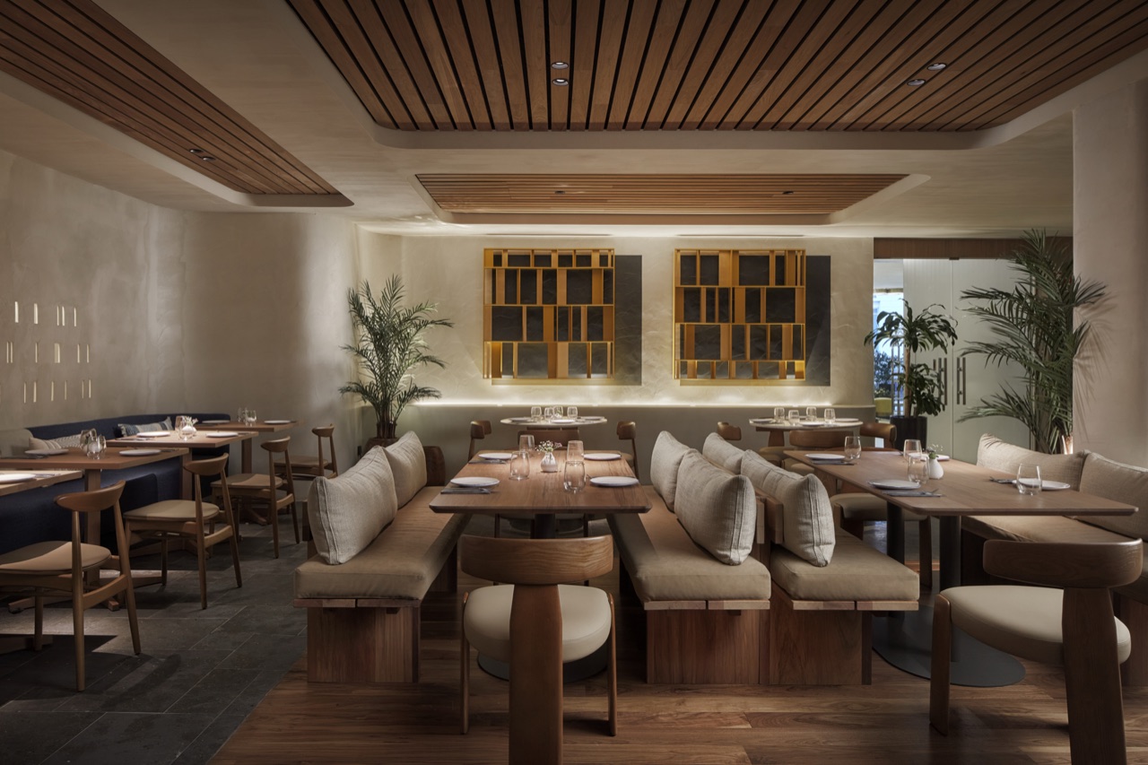 The Tagomago dining room: banquettes with cream cushions, wooden tables set for dinner, a slatted wood ceiling and potted palms.