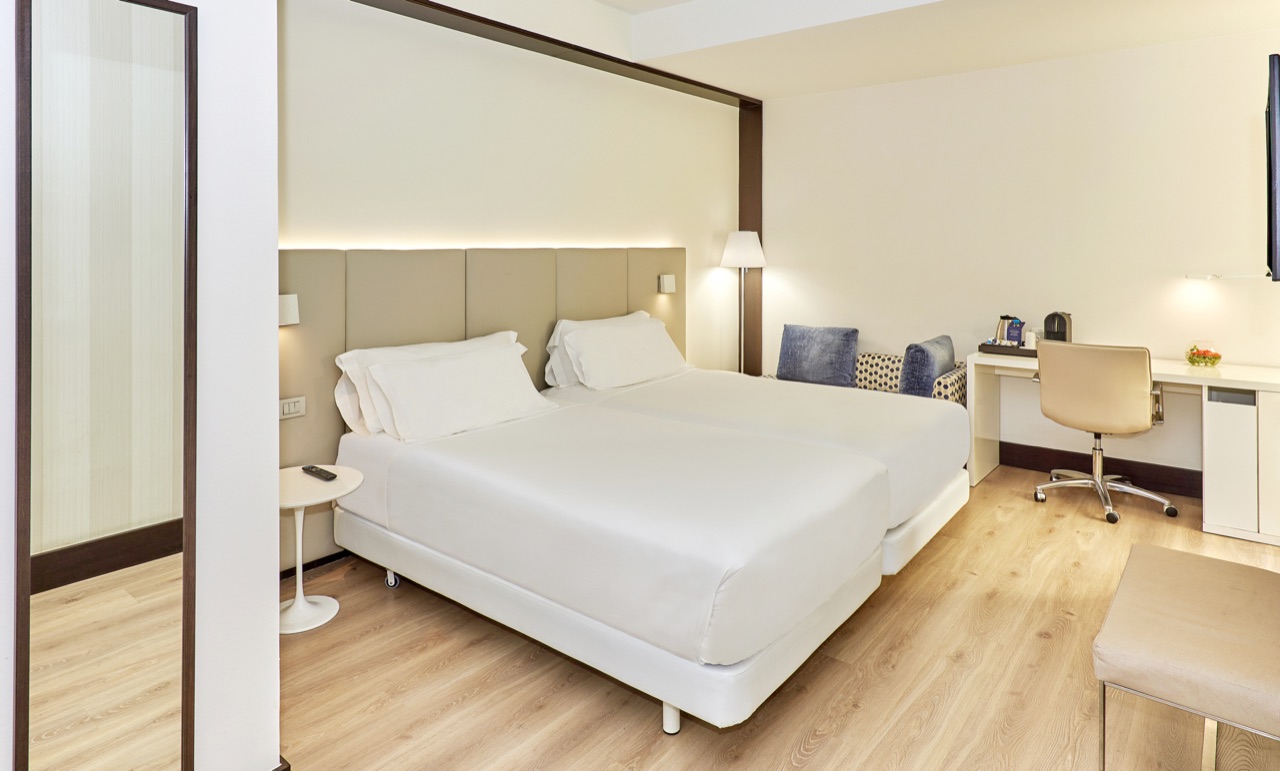 A bright twin room with white bedding, a pale padded headboard wall, light wood floors and a desk by the window.