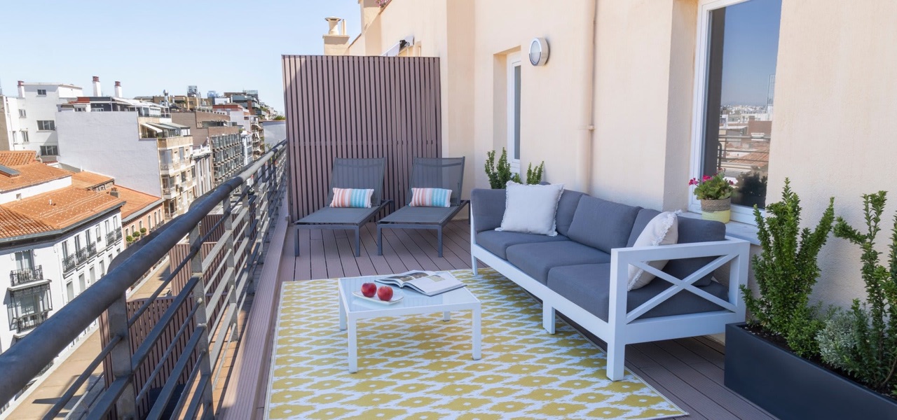A private rooftop terrace with a grey sofa, two lounge chairs, a coffee table, and potted plants overlooking Madrid rooftops.