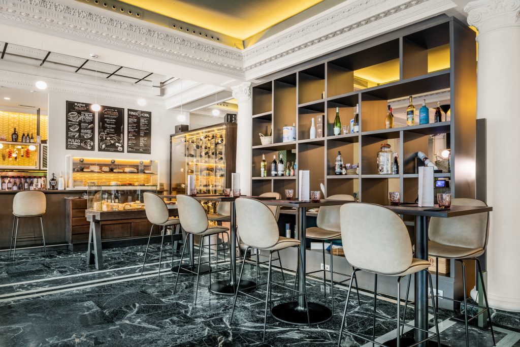 Wine and spirits shelving behind a bar counter with high stools and marble flooring, under an ornate white plaster ceiling.