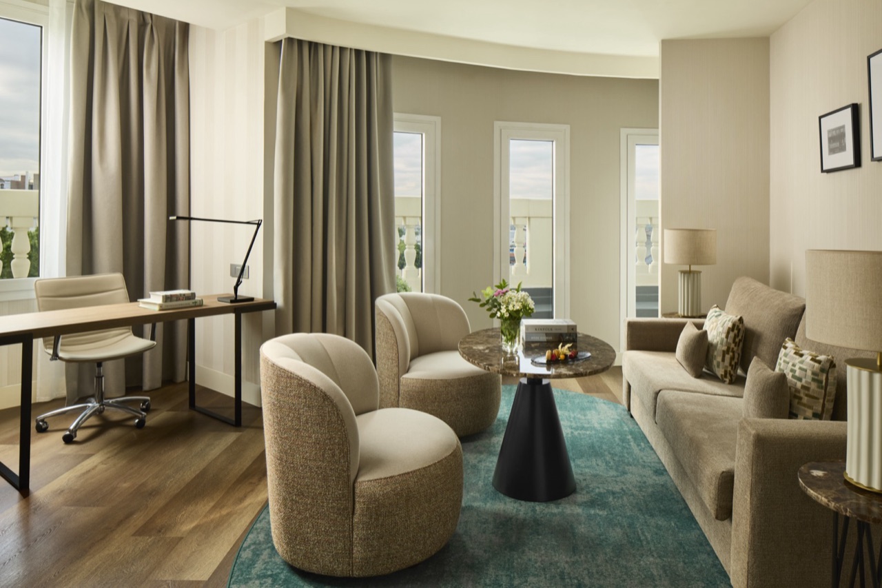 Junior suite sitting area with a desk and chair, two curved armchairs and a sofa around a round marble table, beside curved balcony windows.