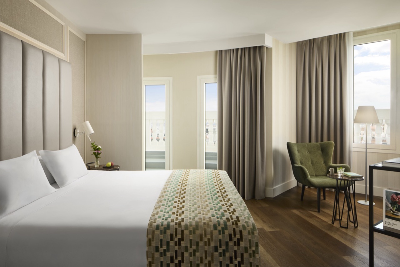 Junior suite bedroom with a channel-tufted grey headboard, white bedding with a woven throw, a green wingback chair and French balcony windows.