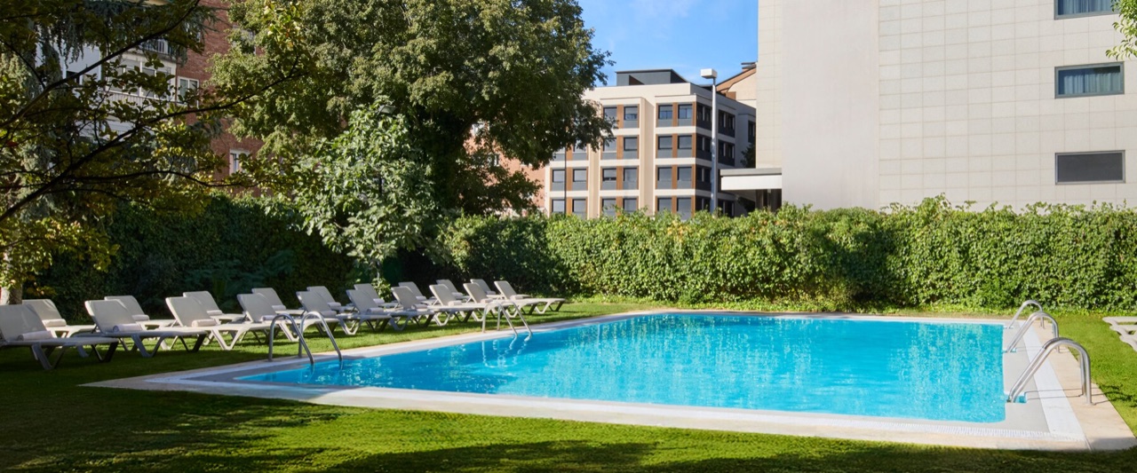 NH Madrid Ventas's outdoor pool set in a lawned garden, lined with white sun loungers and hedges, apartment buildings visible behind.