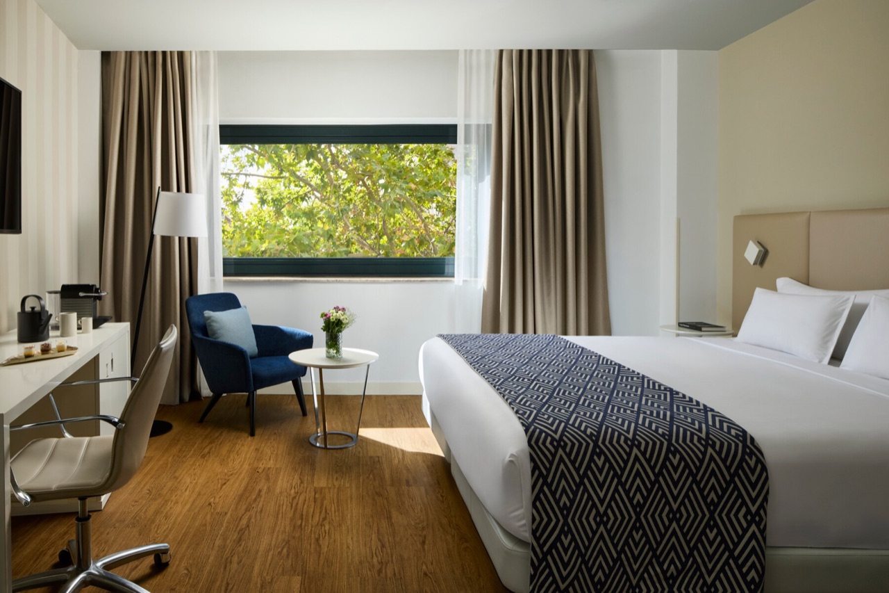 A superior room at NH Madrid Ventas with a king-size bed, a blue armchair by the window and a wooden desk with a coffee machine.