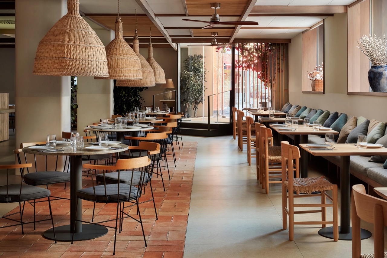 Saona restaurant dining room with wicker pendant lampshades over round marble tables, wooden chairs, a terracotta-tiled floor and a banquette seating area with cushions.