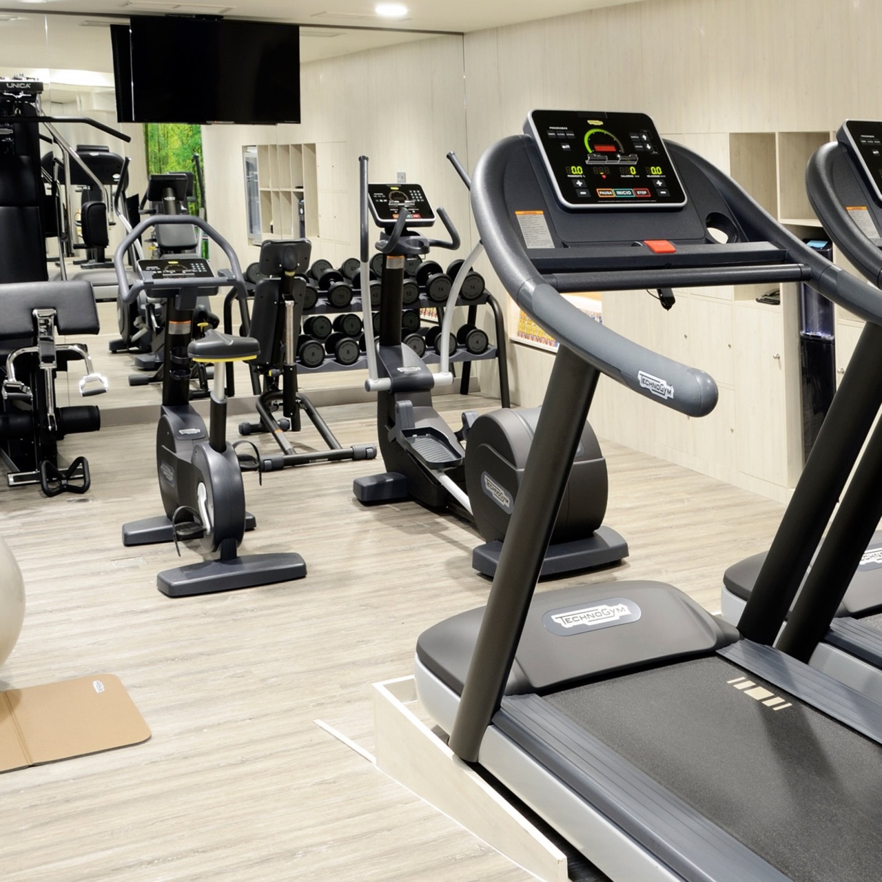 Hotel gym with treadmills, an elliptical machine, exercise bikes and a rack of free weights reflected in a mirrored wall.