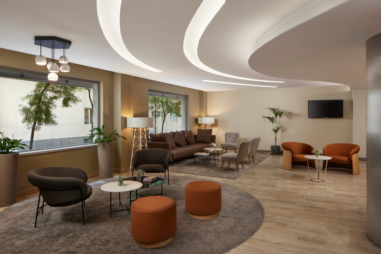 Hotel lounge area with brown, grey and rust-coloured armchairs and sofas grouped on rugs, floor lamps, potted plants and large windows to the street.