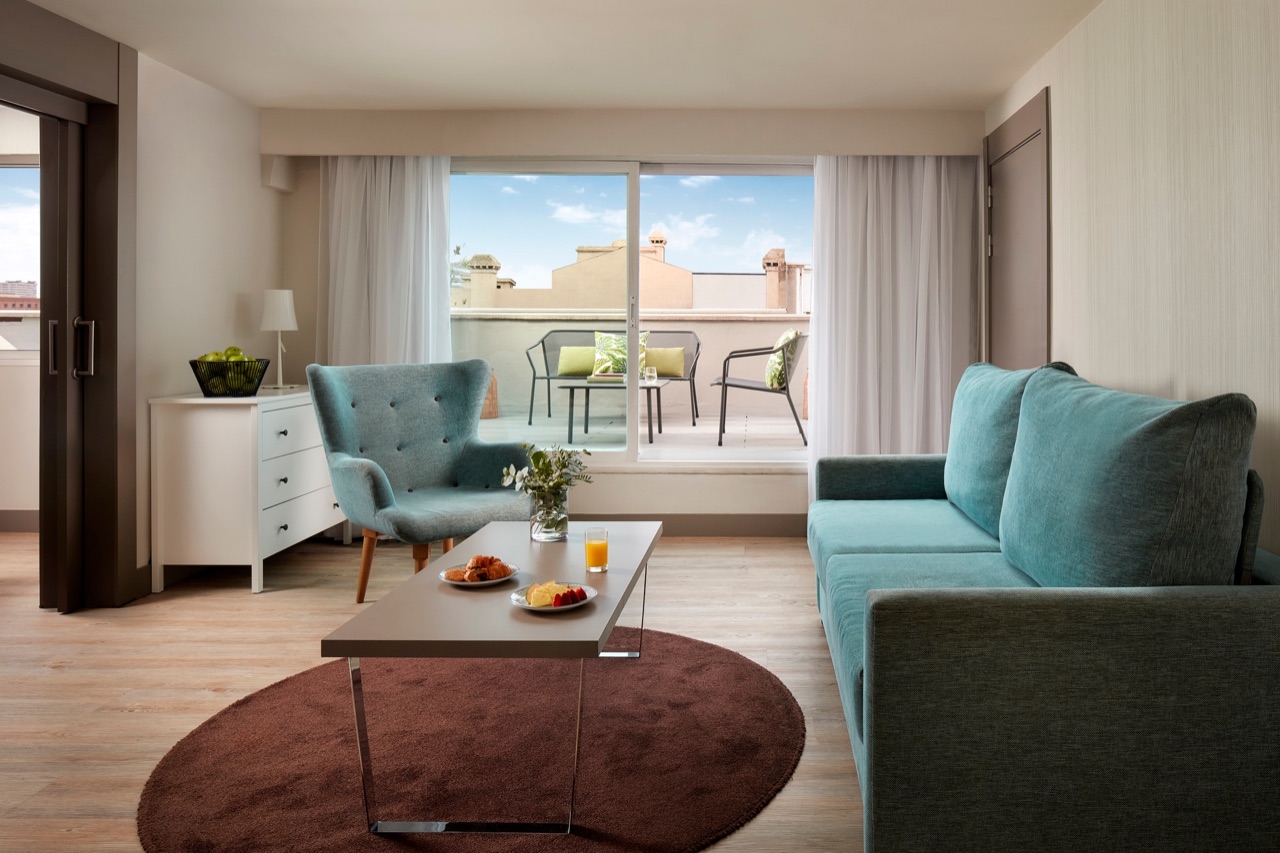 Hotel suite living room with a teal sofa and armchair, a coffee table set with fruit and juice, and a sliding door open to a private terrace with outdoor seating.