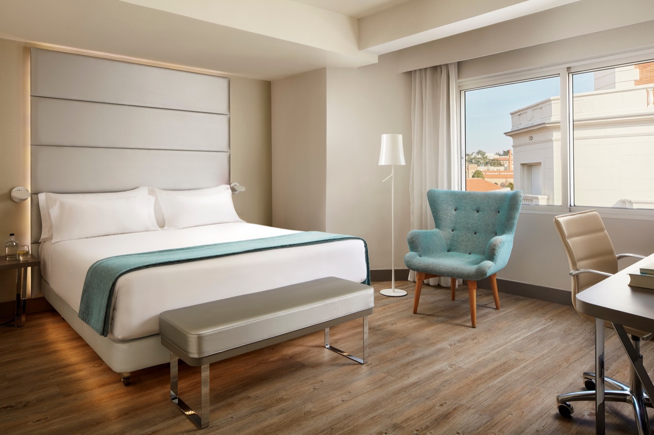 Hotel bedroom with a white bed and teal throw against a tall upholstered grey headboard, a teal armchair and a desk with an office chair by the window.