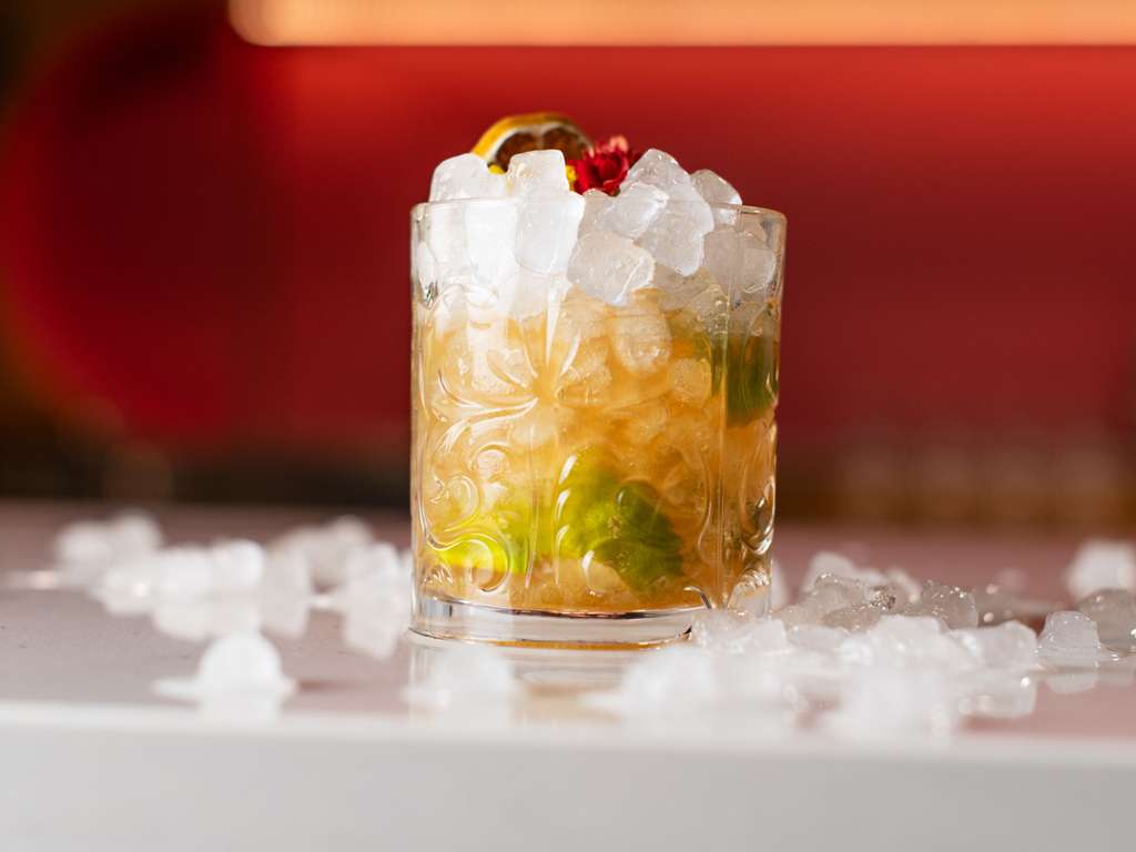 Close-up of an iced cocktail garnished with citrus and flowers on a bar counter with a red-lit background.