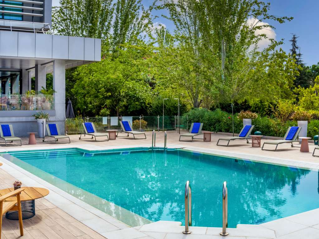 Outdoor swimming pool with blue-cushioned loungers along the deck and the hotel's glass-and-concrete facade behind trees.