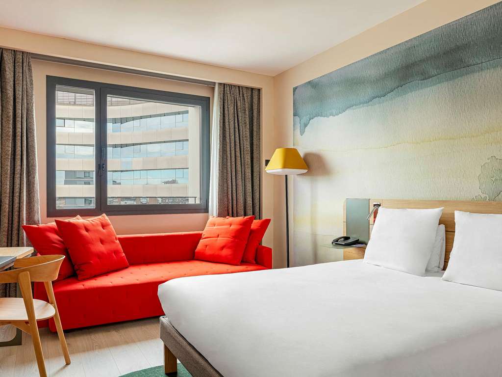 Novotel guest room with a white bed, red upholstered sofa bench and a window overlooking nearby office buildings.