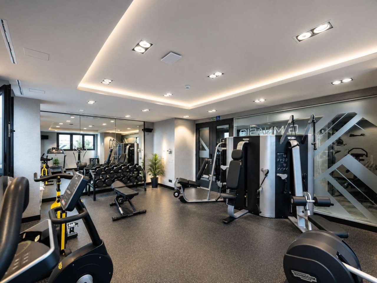 A fitness centre with treadmills and exercise bikes in the foreground, a rack of dumbbells, a weight bench, and cable weight machines along a mirrored wall.