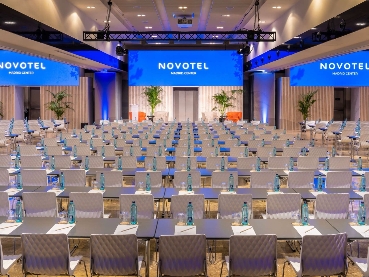 A large conference hall set classroom-style with rows of chairs and desks facing a stage, water bottles and notepads at each seat, and 'Novotel Madrid Center' displayed on screens above the stage.