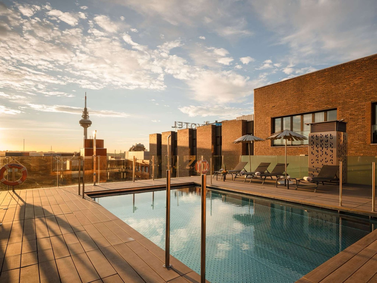 A rooftop swimming pool at sunset with sun loungers and umbrellas on a wood-look deck, a brick hotel building with a Novotel sign, and the Torrespana communications tower visible on the skyline.