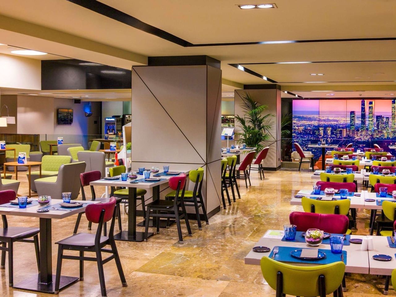 A restaurant dining area with tables set for breakfast, green and dark pink upholstered chairs, a potted plant, and a large backlit photo mural of the Madrid skyline at night.