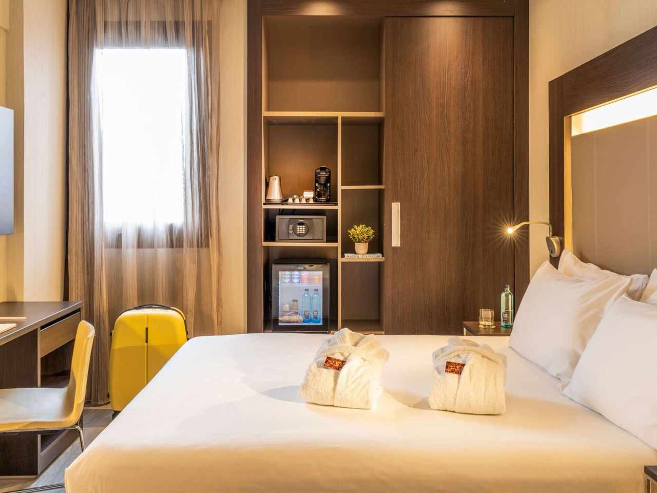 A hotel room with two folded white robes on the bed, a glass-fronted minibar fridge, a wall safe, kettle and coffee machine on open wooden shelving, and a yellow suitcase by the curtained window.