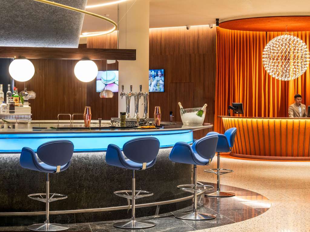 Curved hotel lobby bar with blue upholstered stools, tap handles and an orange reception desk in the background.