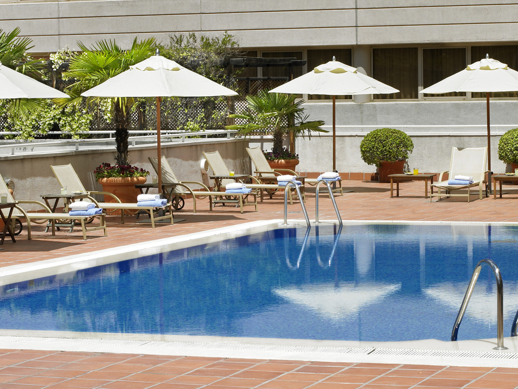 Outdoor swimming pool lined with sun loungers, white umbrellas and potted palms.