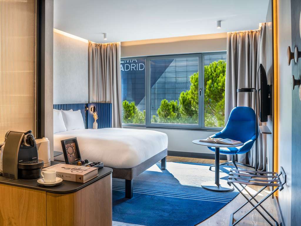 Hotel bedroom with a white bed, blue armchair and a window looking out at the IFEMA Madrid building.