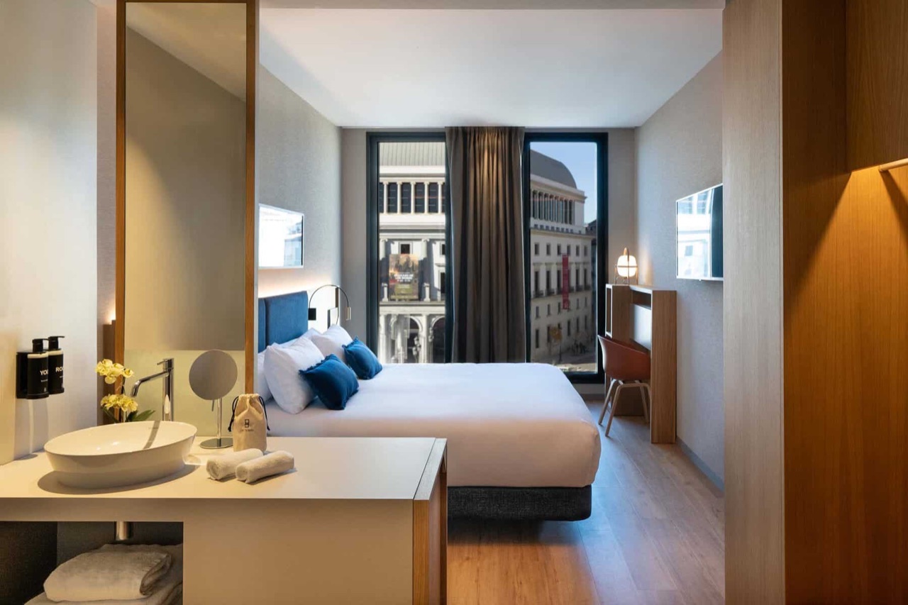 A hotel room with a blue upholstered headboard, white bedding and floor to ceiling windows looking out at the Teatro Real building across the plaza.