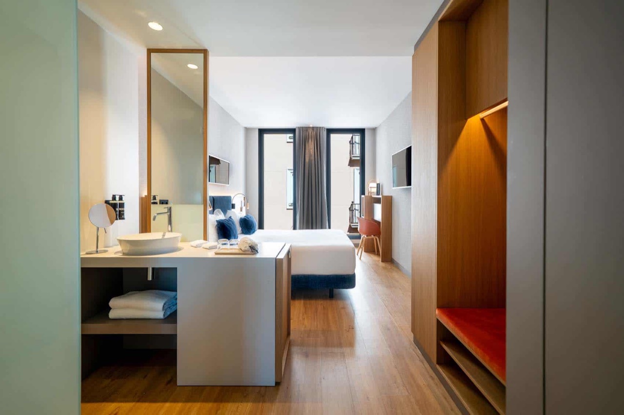 A hotel room with a white bed, an open bathroom vanity with a round sink and mirror in the foreground, and a wall mounted television beside the window.