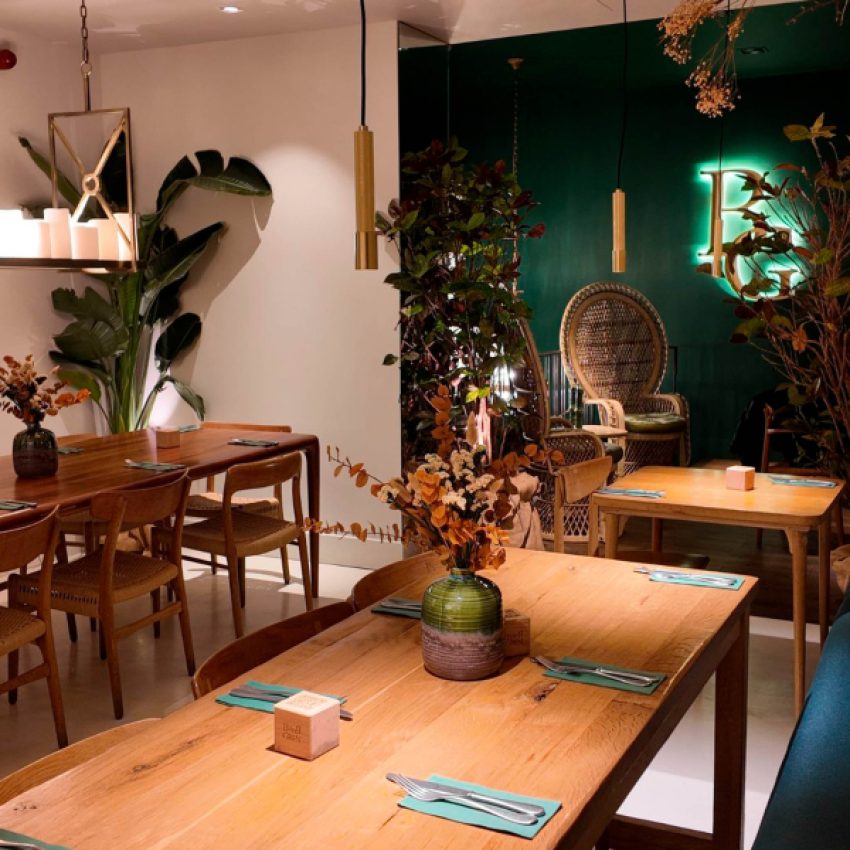 The Bumpgreen dining room with wooden tables set for service, rattan peacock chairs and a dark green wall with an illuminated BG logo.