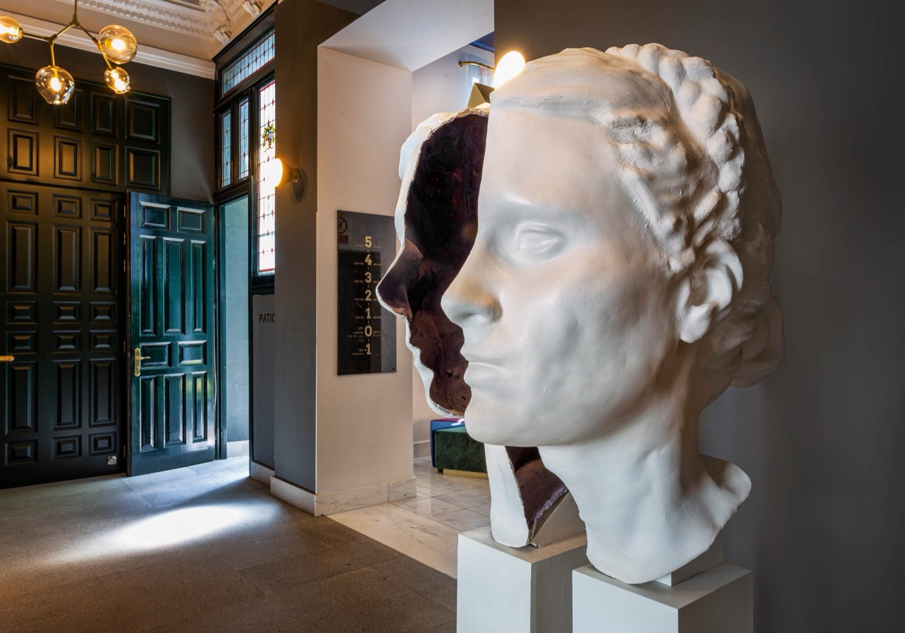 A dark lobby corridor with a split-face plaster head sculpture on a plinth, stained-glass windows and a dark green panelled door.