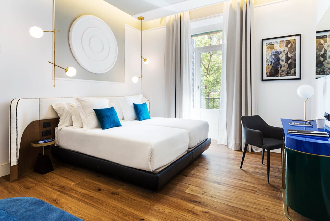 A twin-bed room with blue velvet cushions, a wood-panelled headboard, brass reading lamps and a balcony door open to greenery outside.