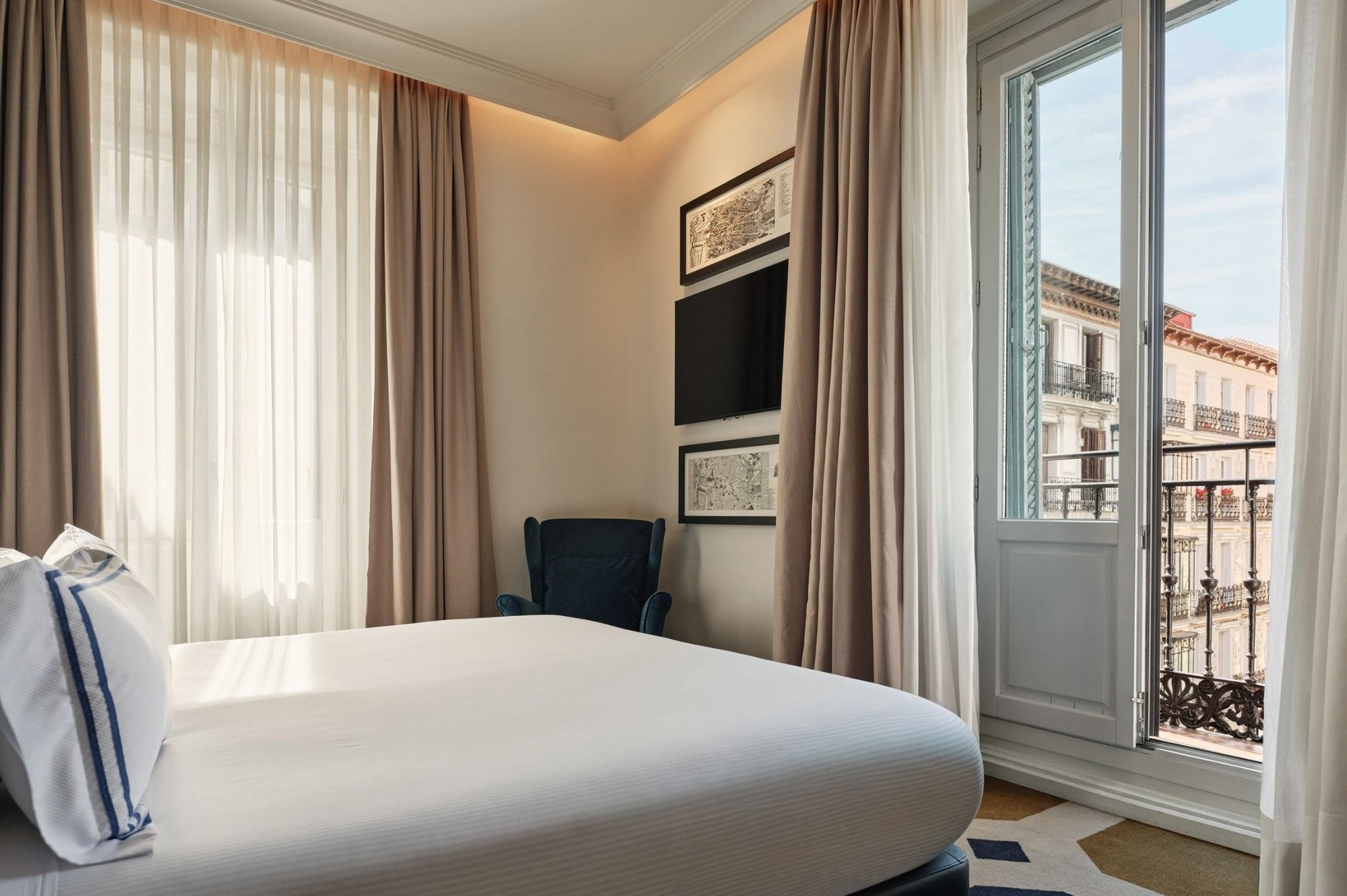 A bedroom with a double bed, an armchair and a French window with a balcony looking onto neighbouring buildings.