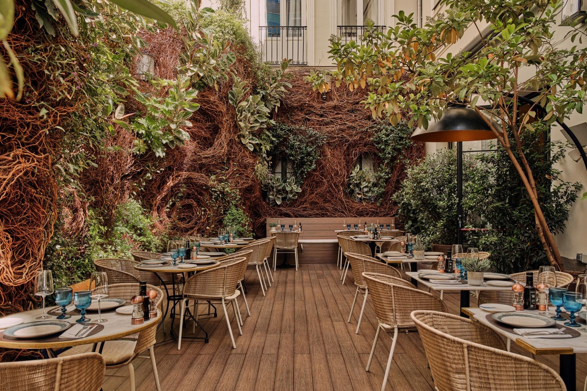 An outdoor terrace in a courtyard with a planted wall, trees and wooden decking, set with café tables and wicker chairs.