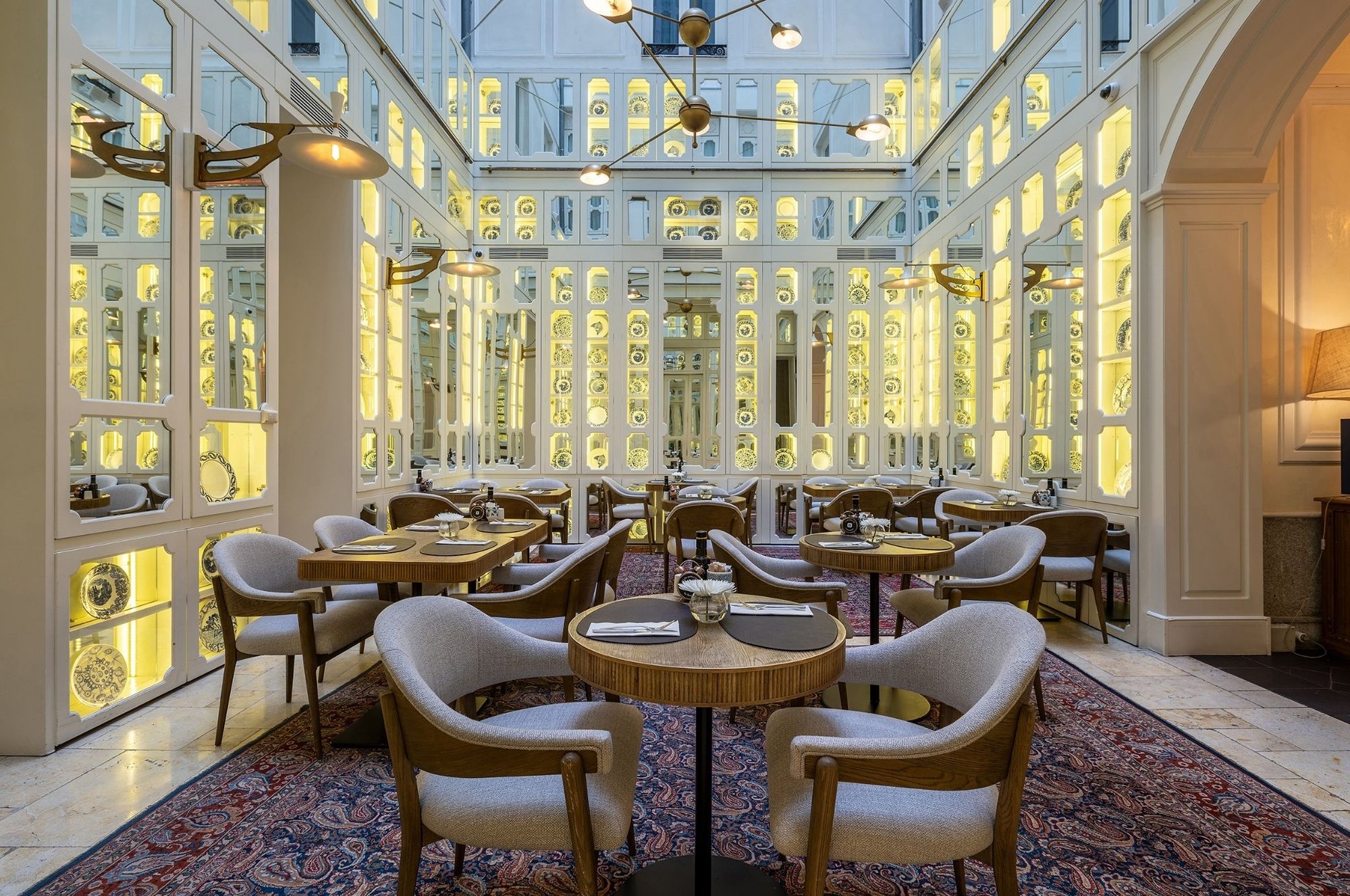 A courtyard dining room with round wooden tables and upholstered chairs on a patterned rug, under a glass roof with lit white lattice walls.