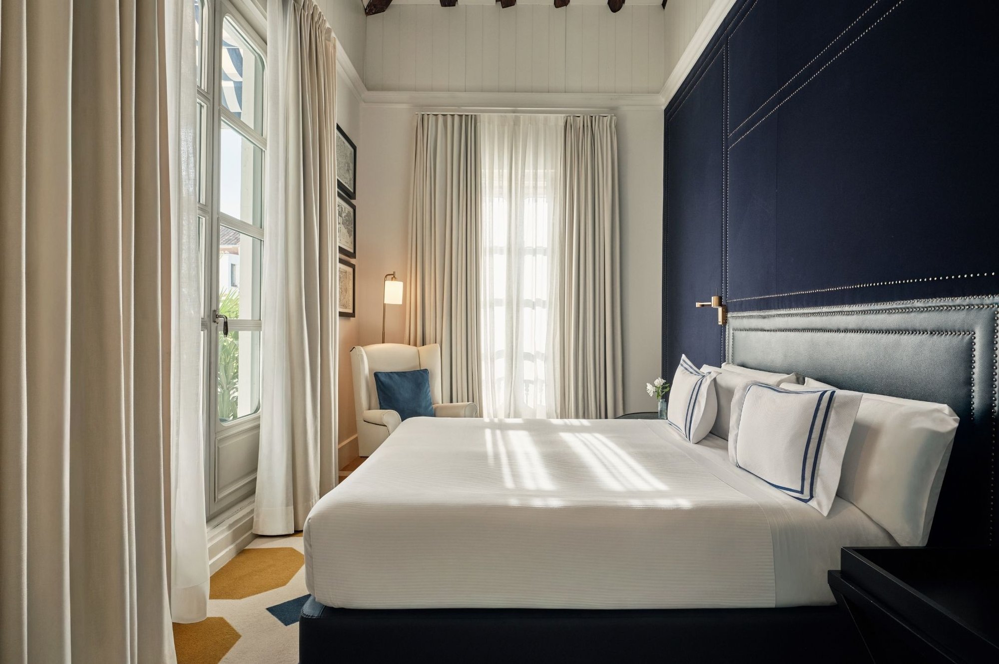 A bedroom with a double bed in white linen against a dark navy panelled headboard wall, tall shuttered windows and a small armchair.