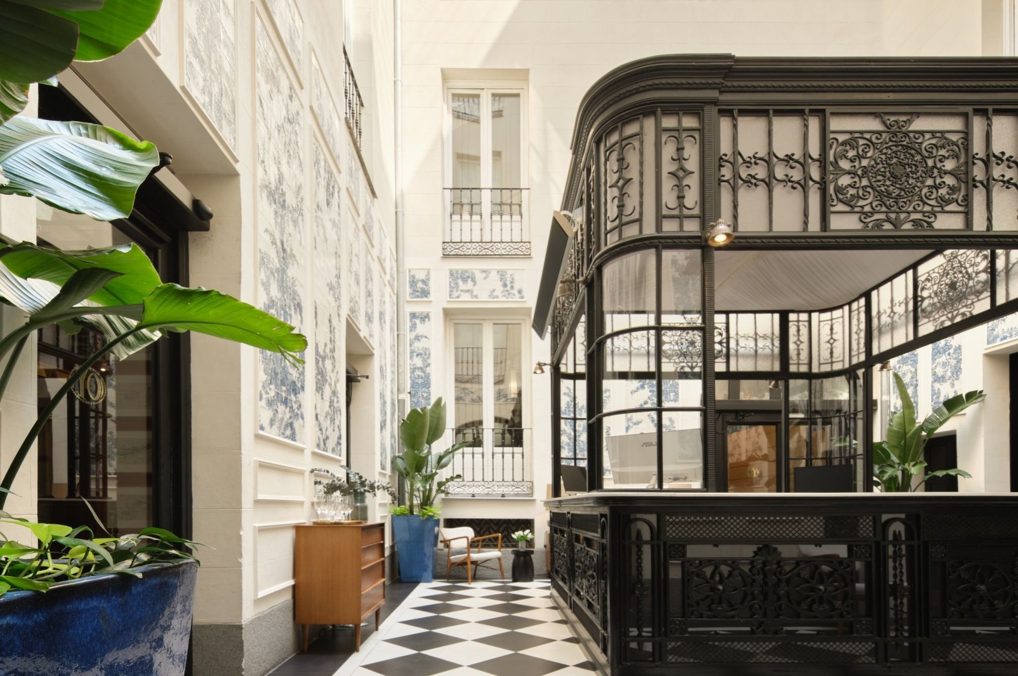 A courtyard with blue-and-white patterned walls, a black wrought-iron glass pavilion, a chequered black and white floor and large green plants.