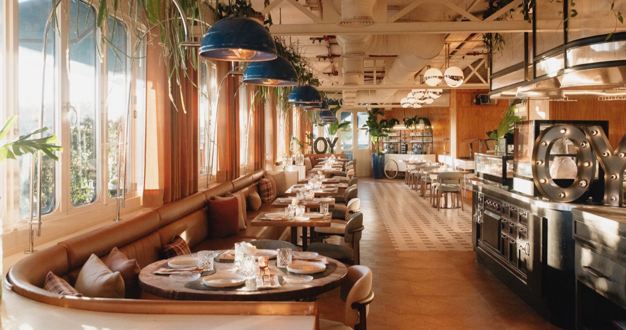 A restaurant dining room with a curved leather banquette, set tables, hanging blue pendant lamps, and a lit 'OY' marquee sign by the counter.