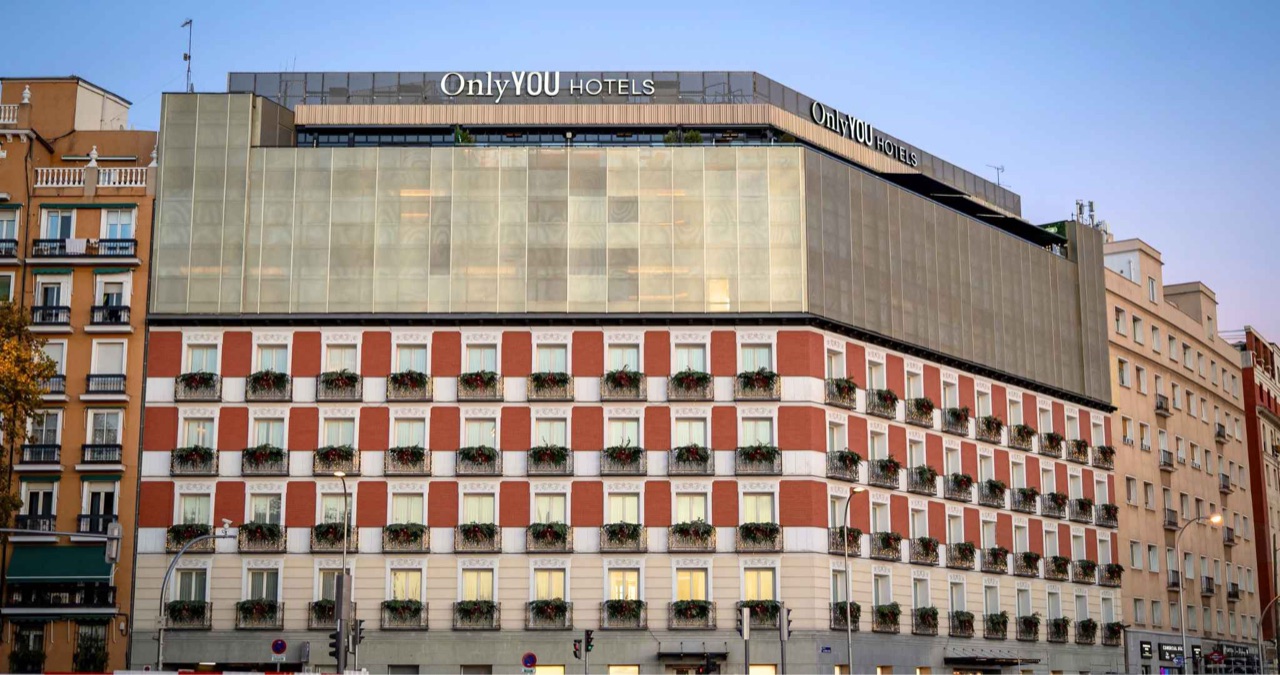 The hotel's restored red-and-white brick facade at dusk, topped by a glass upper floor with an illuminated Only YOU Hotels sign.