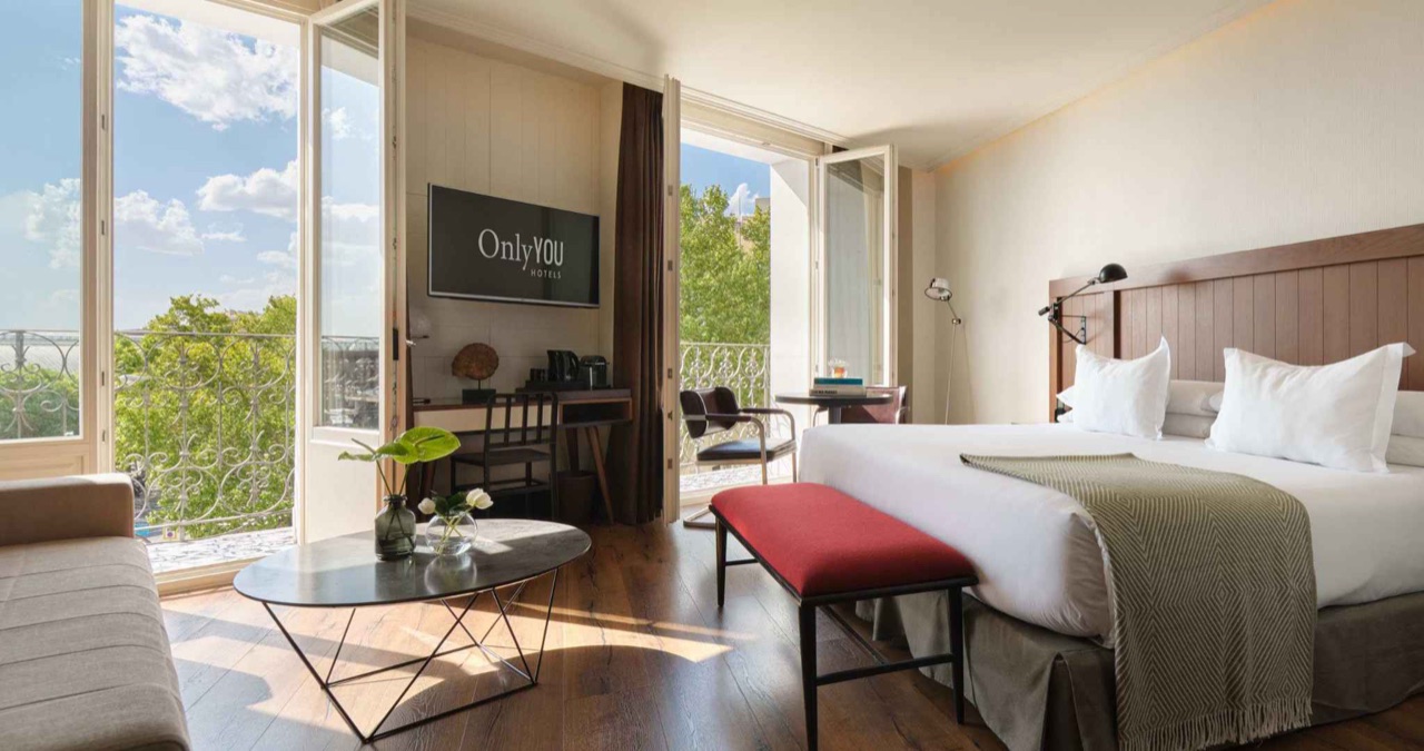 A hotel room with a wood-panelled headboard, a red upholstered bench at the foot of the bed, and open French balcony doors overlooking tree-lined streets.