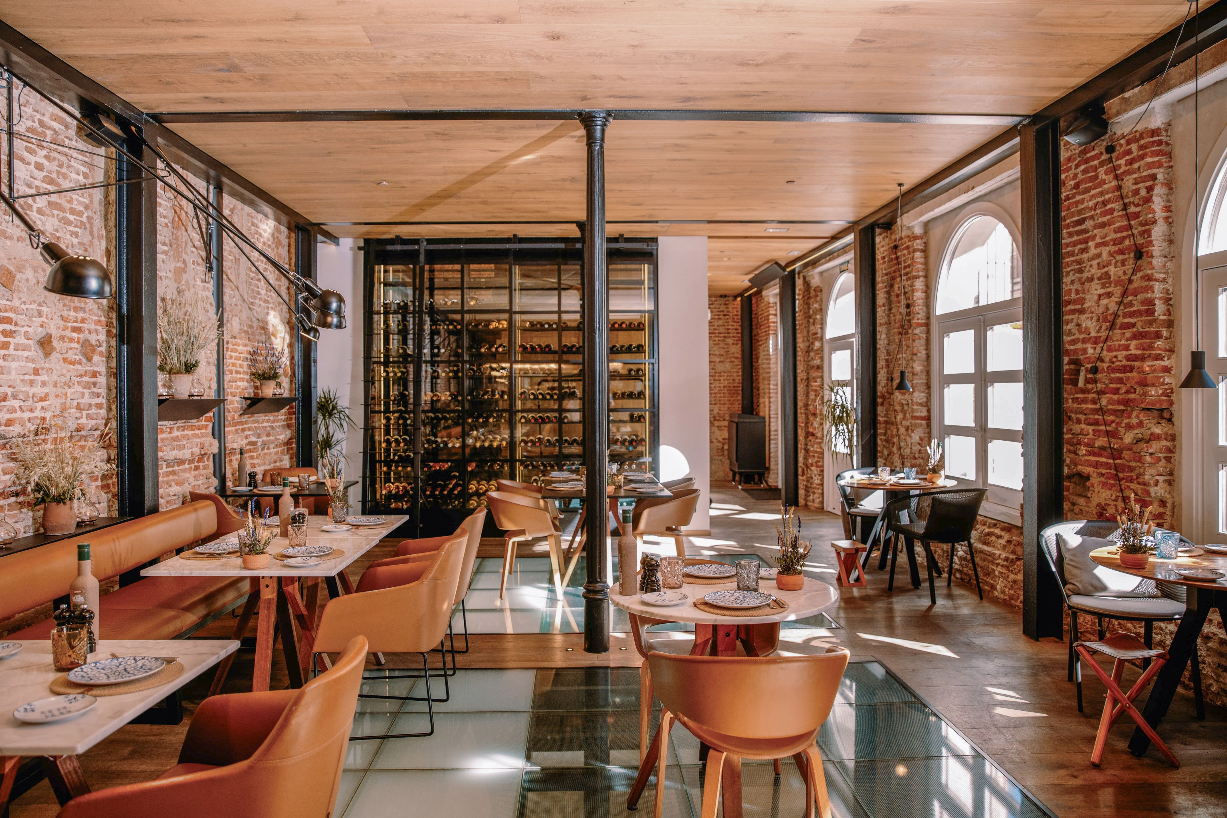 A restaurant with exposed brick walls, a black cast-iron column, a glass-fronted illuminated wine cellar, marble-topped tables with tan leather chairs and inset glass floor panels.