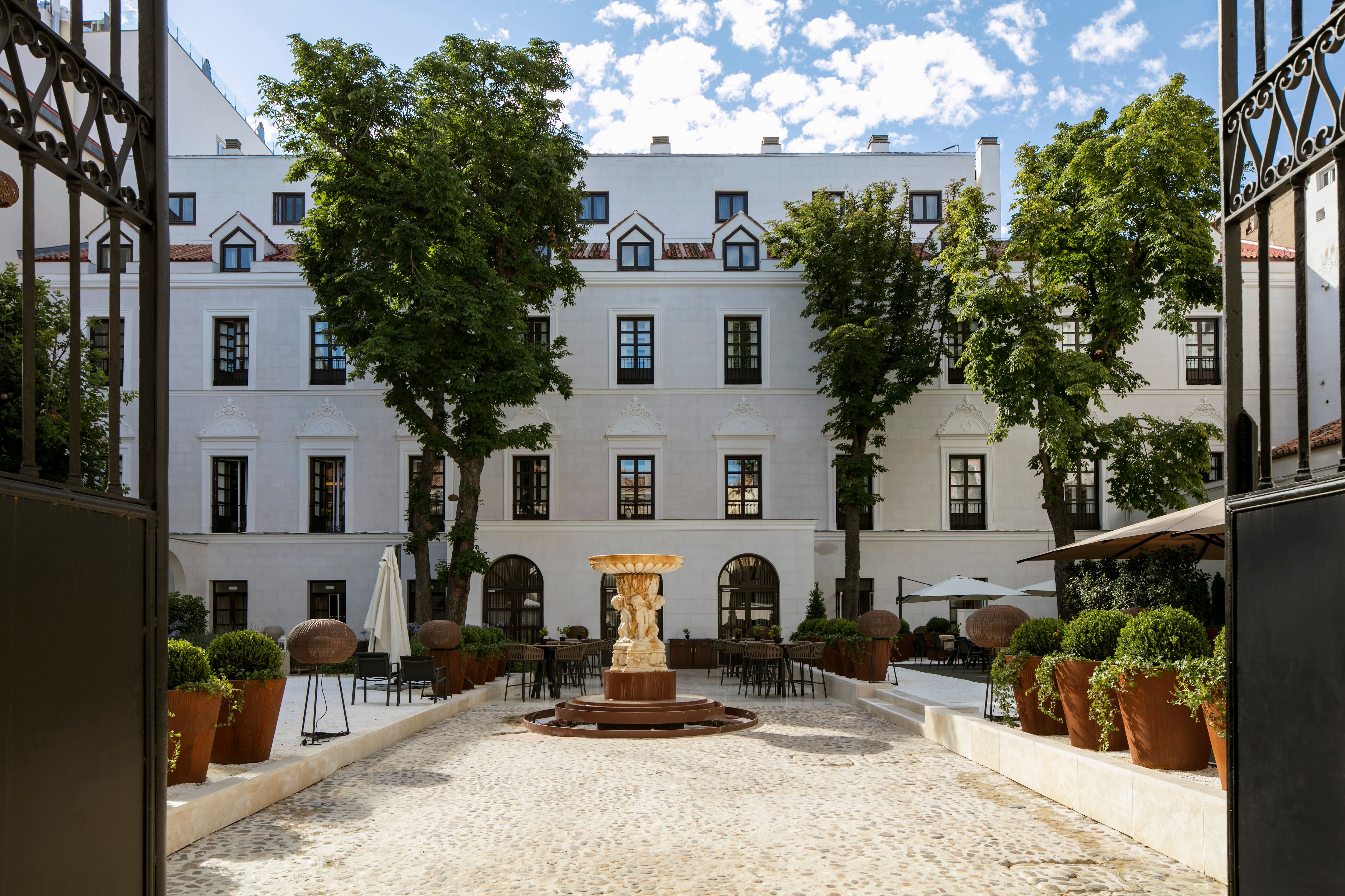 A white four-storey palace facade with dormer windows and carved window pediments seen through open iron gates across a cobbled courtyard with a stone fountain, potted shrubs and outdoor tables under trees.