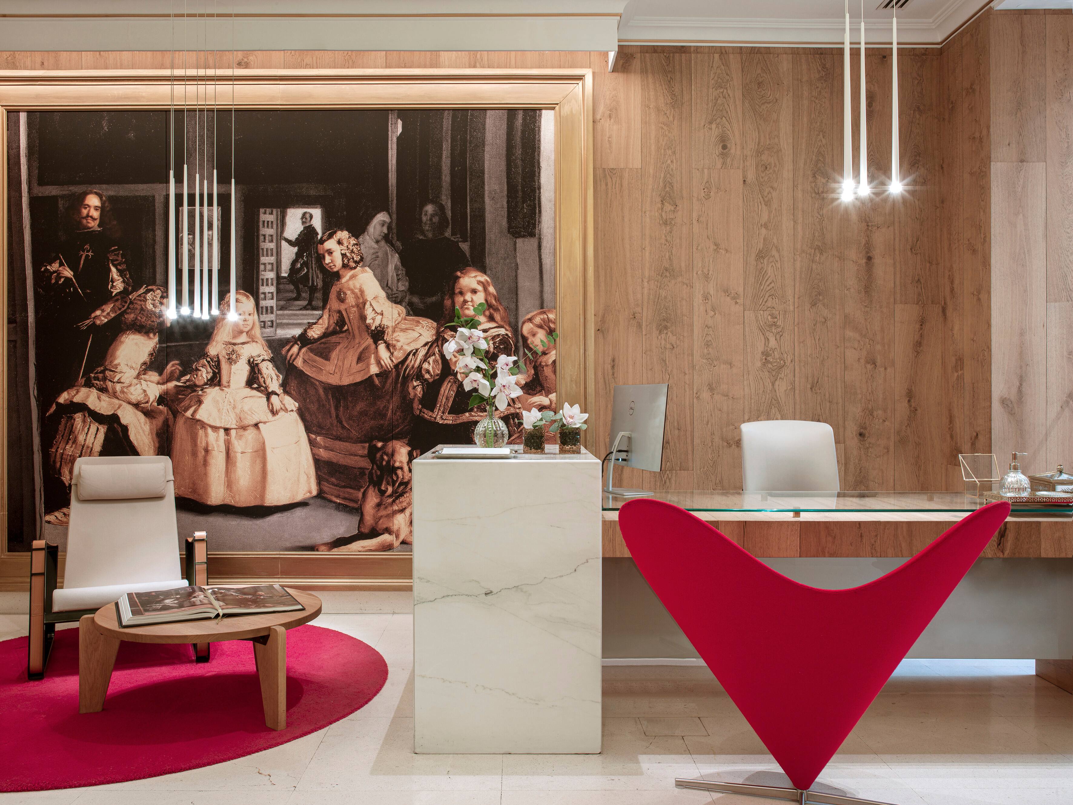 A reception area with a large gold-framed reproduction of Las Meninas on a wood-panelled wall, a white marble desk, a glass-topped counter and a red heart-shaped chair on a magenta rug.