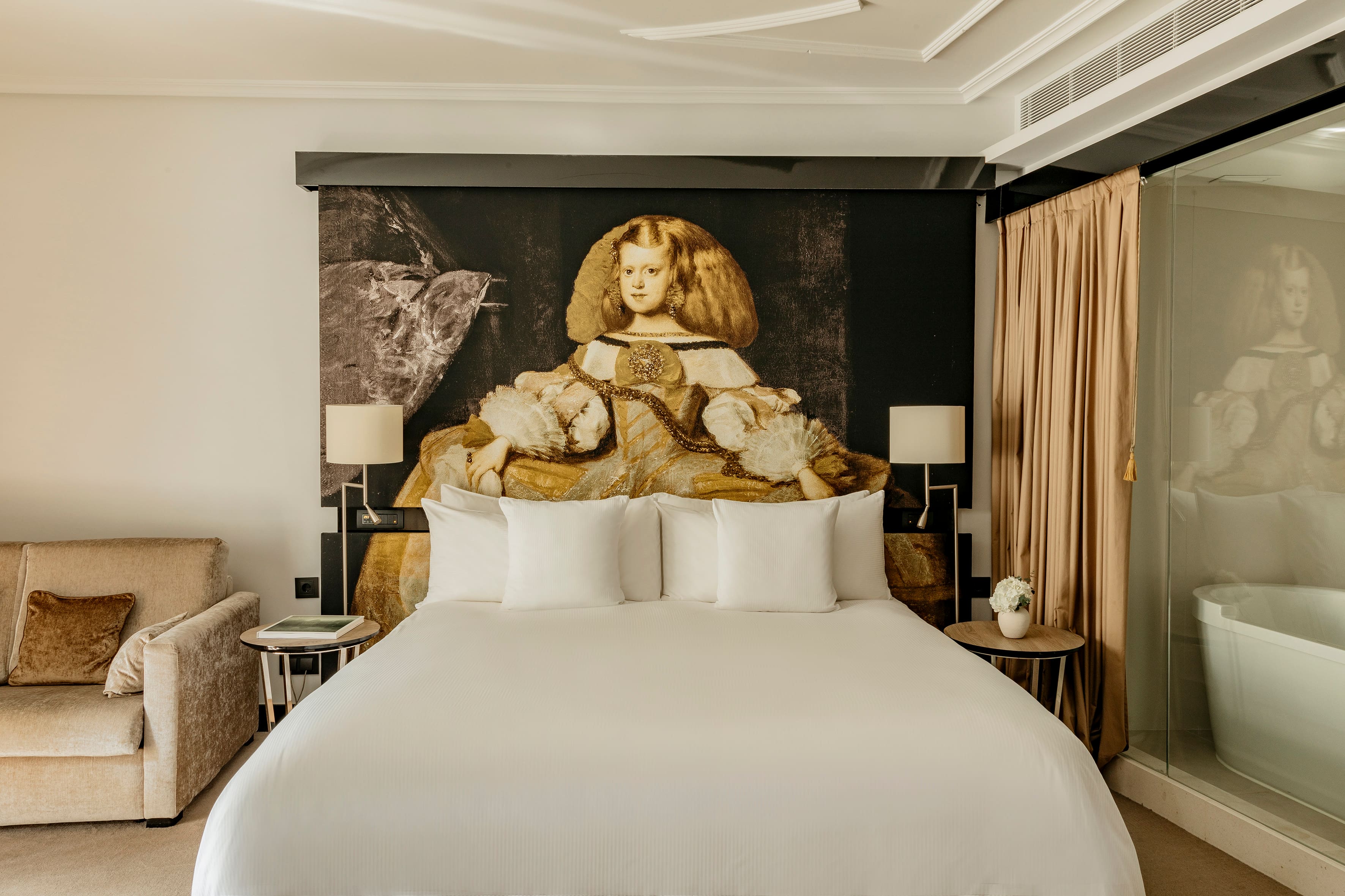 A guest room with a wide bed in white linens beneath a large painted portrait panel headboard, twin shaded lamps on round side tables, a beige armchair and a freestanding bathtub behind a glass partition.