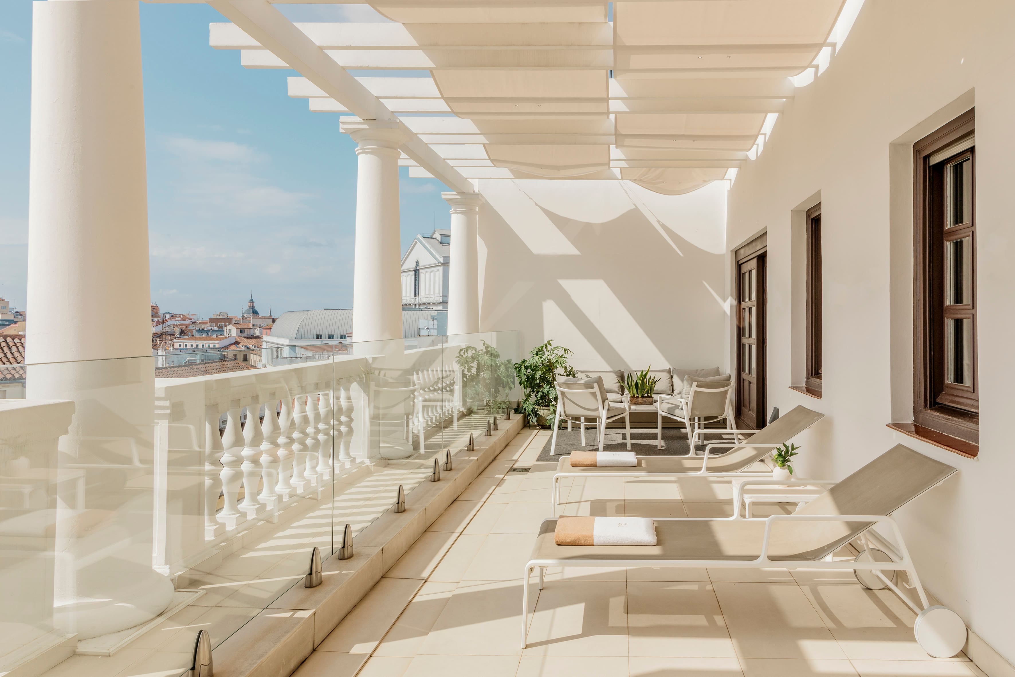 A rooftop terrace with white columns and a slatted pergola with fabric shades, sun loungers and cushioned armchairs along a stone balustrade overlooking tiled rooftops and a distant dome.