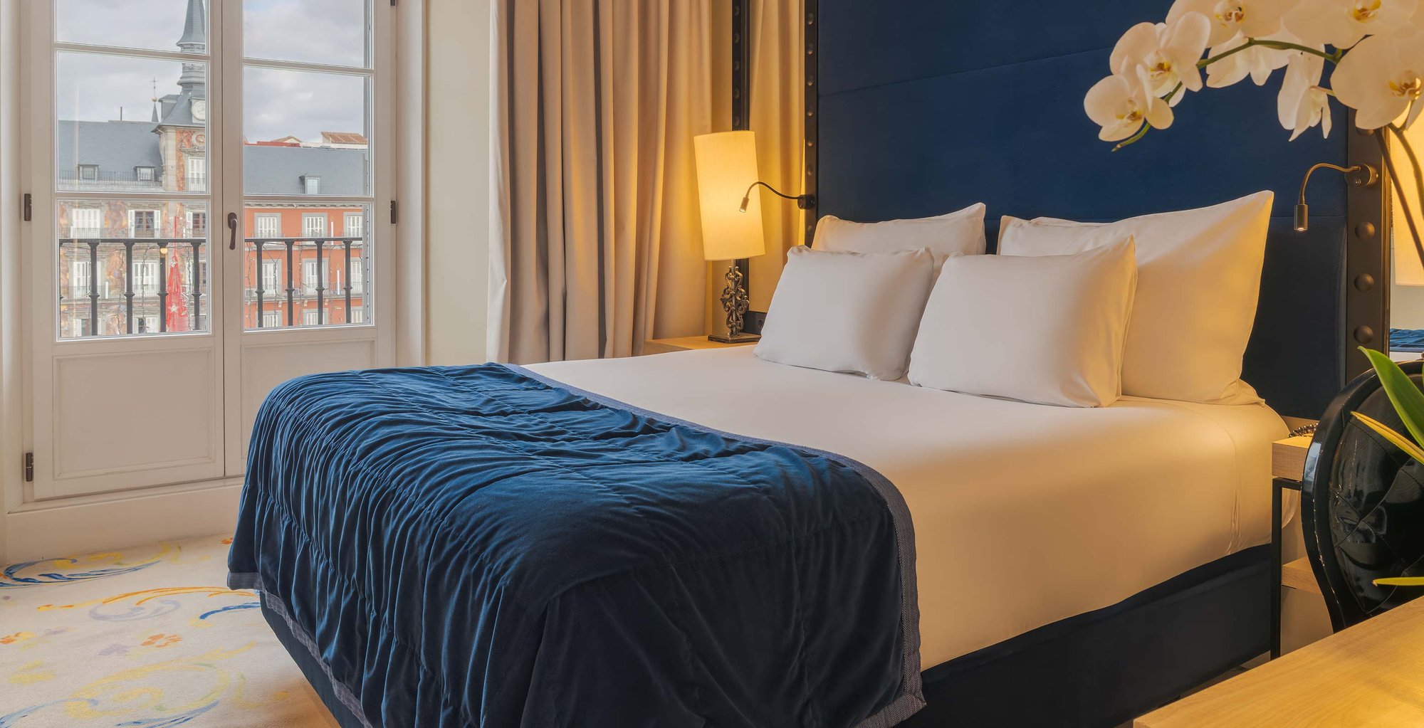 A double bed with a dark blue velvet throw and tall blue headboard, beside a balcony door that looks across to the painted facade of Plaza Mayor.