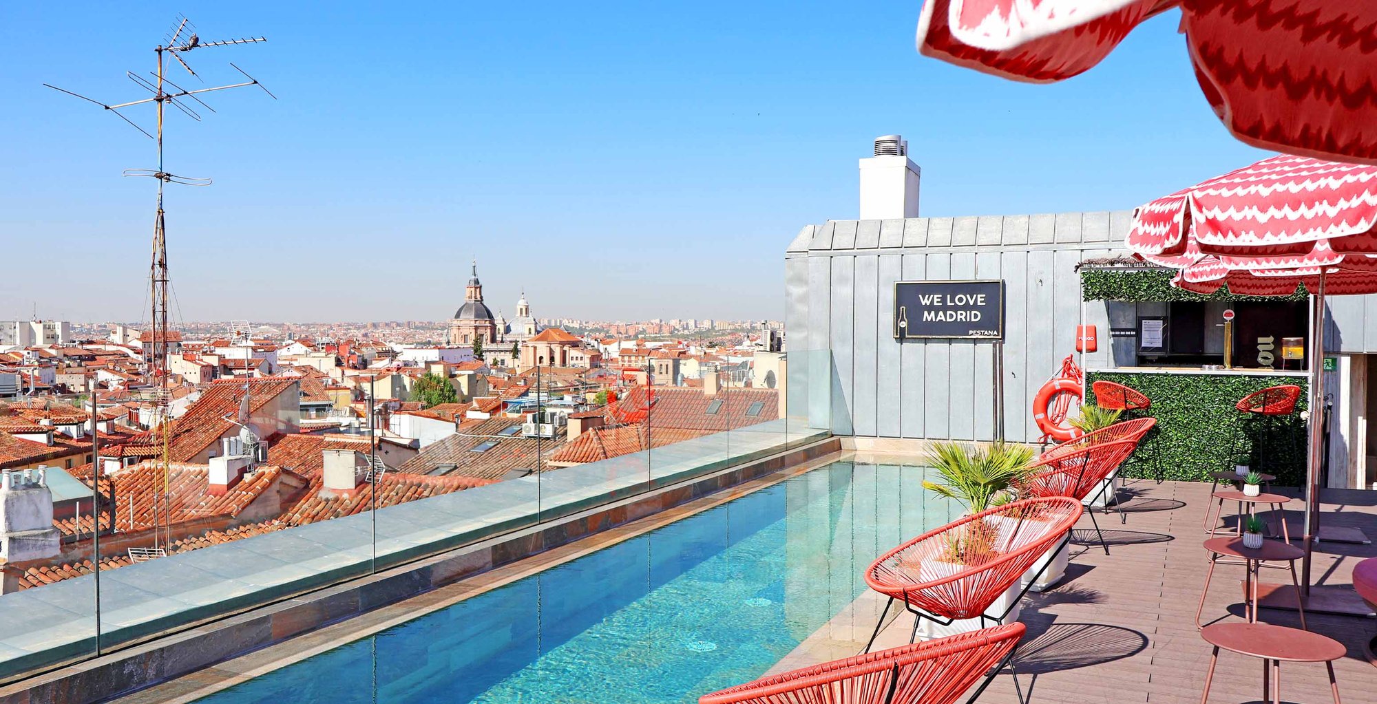 The rooftop pool with a glass balustrade, red wire chairs and parasols, and a view over Madrid's terracotta rooftops to a distant dome.