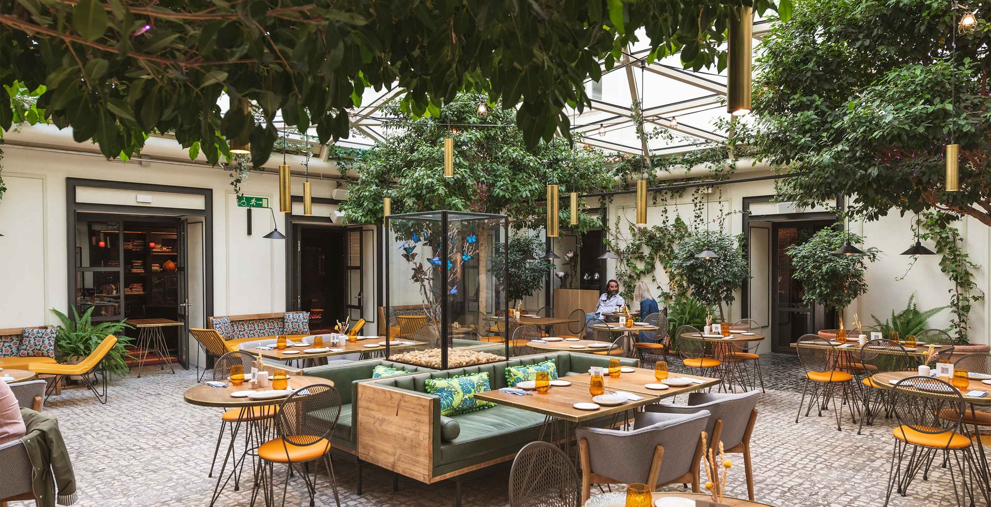 The glass-roofed inner patio restaurant with large leafy trees, wooden tables, green banquettes and orange chairs on a cobbled floor.