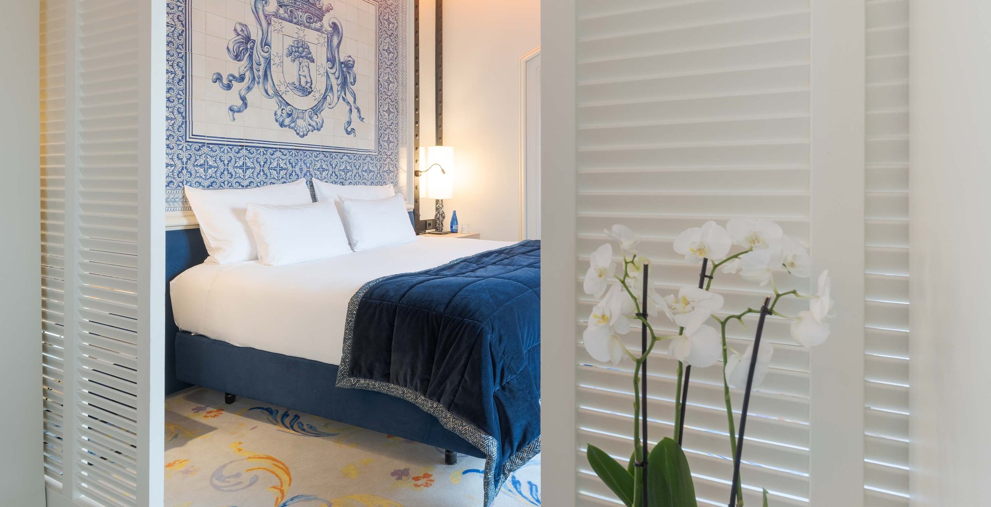 A bed with a blue velvet throw set against a blue-and-white tile panel showing a crowned coat of arms, seen between white louvred shutters.