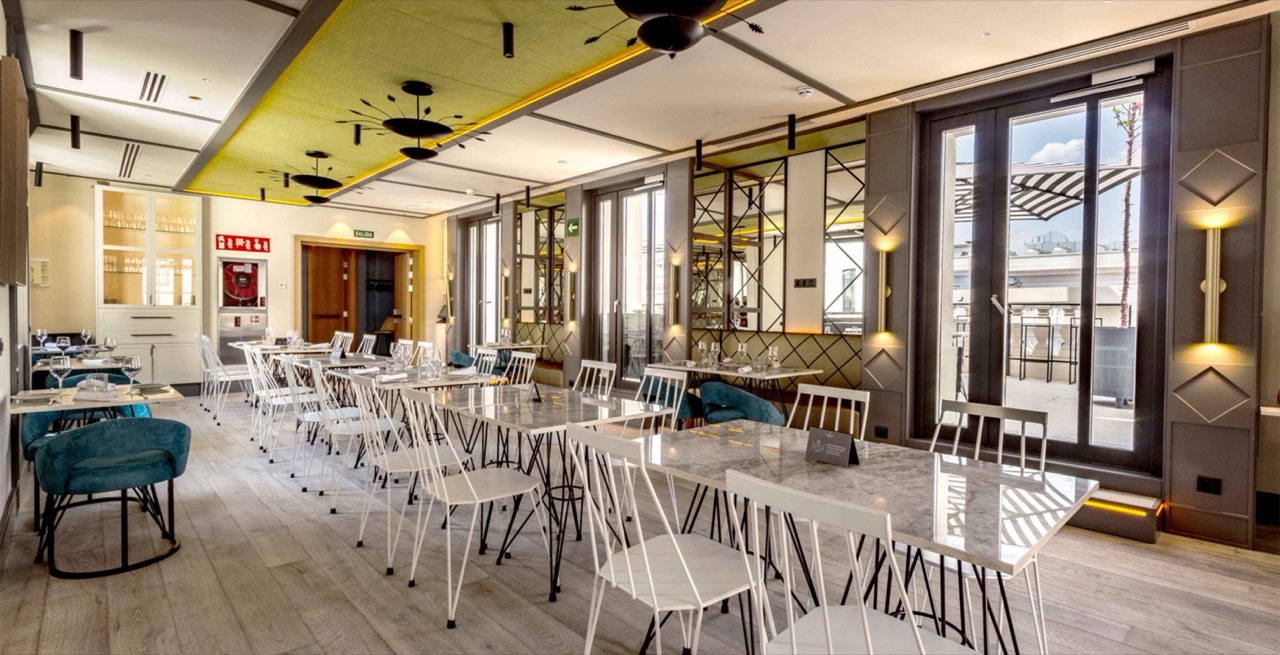 Empty 9th-floor pizzeria dining room with long marble-topped tables, white chairs, and glass doors opening onto a rooftop terrace.
